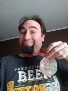 Man with a goofy smile holding a cardboard medallion reading "World's Best Dad"